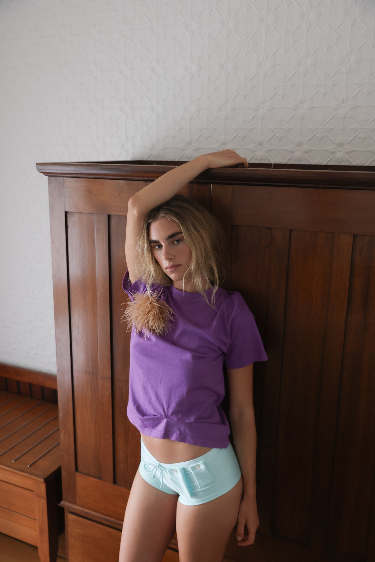 A woman wearing a purple short-sleeve t-shirt and aqua shorts stands against a dark wooden headboard in a bedroom setting.