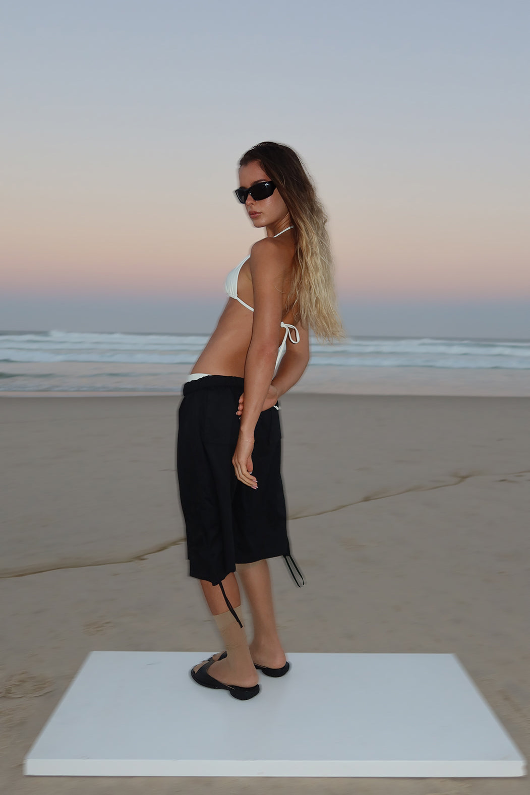 Woman in a cream bikini top and black shorts standing on a beach at sunset.